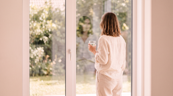 Woman looking out a bright window holding a glass of water, loving someone you cannot talk about