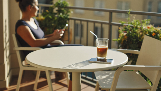 Sunlit balcony table with an empty chair opposite, a half-finished iced coffee and a phone, the waiting inside a situationship hope cycle