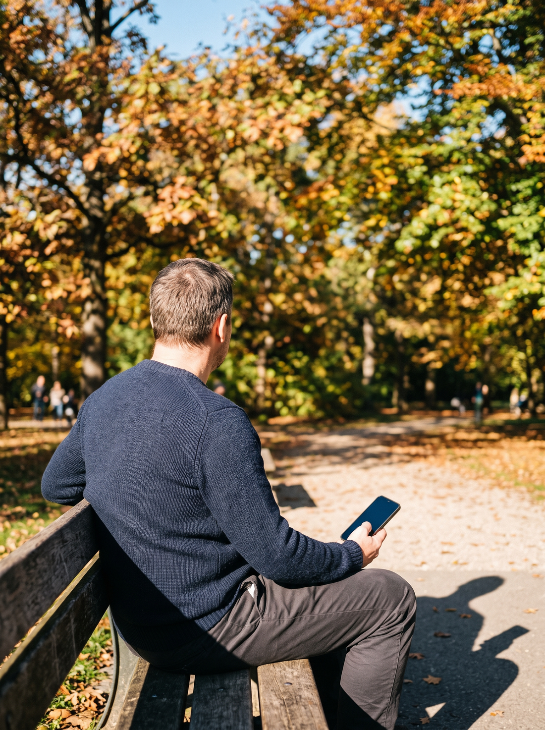 Seen from behind, a man sits on a park bench under autumn trees, a phone in his hand